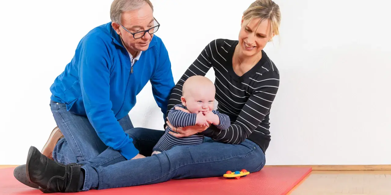 Ein Mann und eine Frau üben mit einem Baby auf einer roten Matte. Das Baby greift nach seinen Fingern. kg zns kinder.