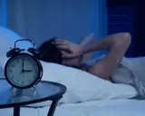 Eine Frau hält sich im Bett die Ohren zu, während ein Wecker auf 3 Uhr steht. Warum wache ich immer um 3 Uhr auf?