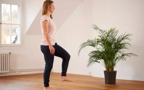 Frau übt auf einem Bein stehen im hellen Raum, neben einer grünen Palme.