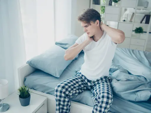 Junger Mann sitzt auf dem Bett, reibt sich den Nacken. Er hat nackenschmerzen nach dem schlafen.