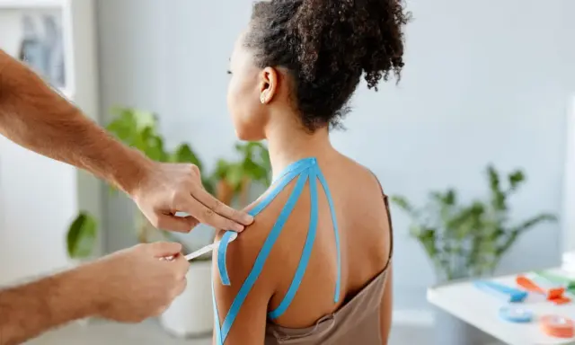 Therapeut legt blaues Kinesio-Tape an Schulter an. Vielleicht ein neues, modernes "Omas Hausmittel gegen Schulterschmerzen"?