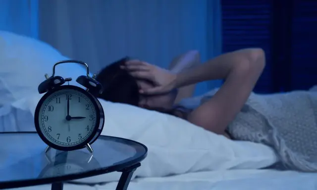 Eine Frau hält sich im Bett die Ohren zu, während ein Wecker auf 3 Uhr steht. Warum wache ich immer um 3 Uhr auf?