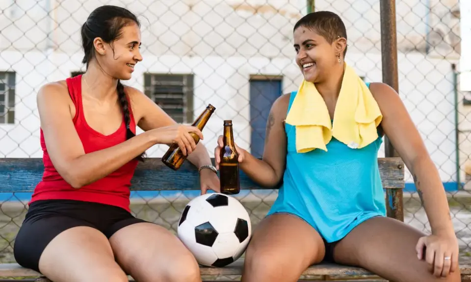 Zwei Frauen mit Fußball und Bierflaschen stoßen an. Nach dem Sport ist vor dem Genuss: sport und alkohol.