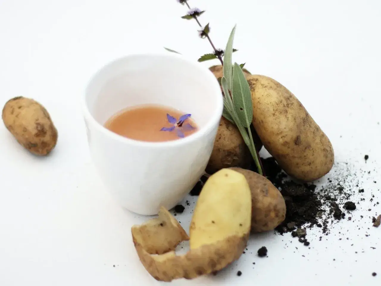 Ein weißer Becher mit Kartoffelsaft, garniert mit einer Blüte. Kartoffeln und Kräuter daneben. Hilft bei Sodbrennen.