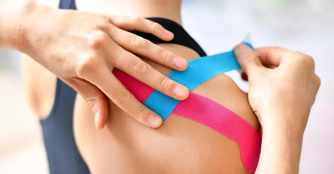 H&auml;nde legen pinkes und blaues Tape auf eine Schulter. Ein modernes Mittel gegen Schulterschmerzen, das an Omas Hausmittel erinnert.