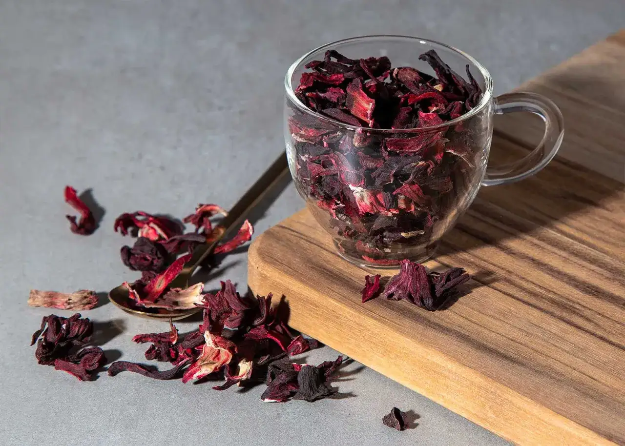 Get ready for the Cistus Tee Wirkung! A glass cup overflows with dried hibiscus petals, a golden spoon rests nearby, hinting at the refreshing taste and health benefits.