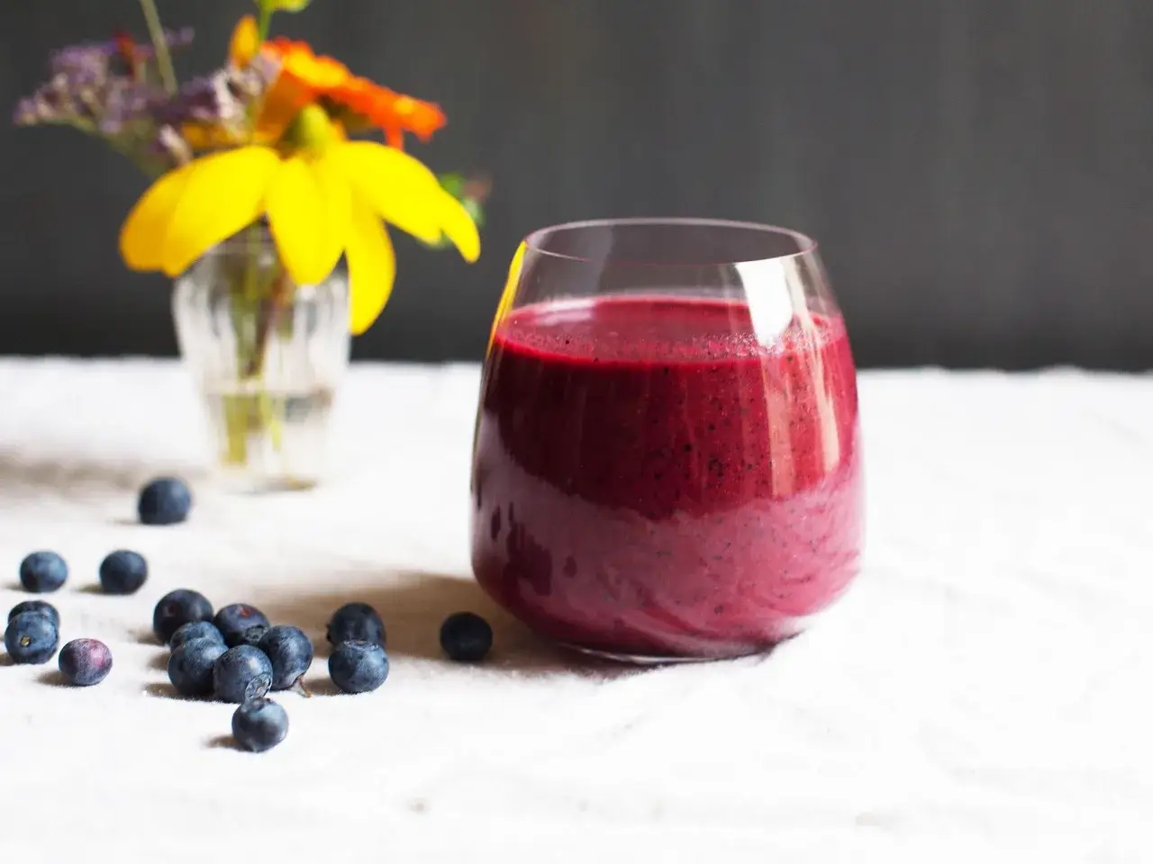 Ein Glas leuchtend roter Smoothie mit rote Bete und Blaubeeren, daneben frische Beeren und ein kleiner Blumenstrau&szlig;.