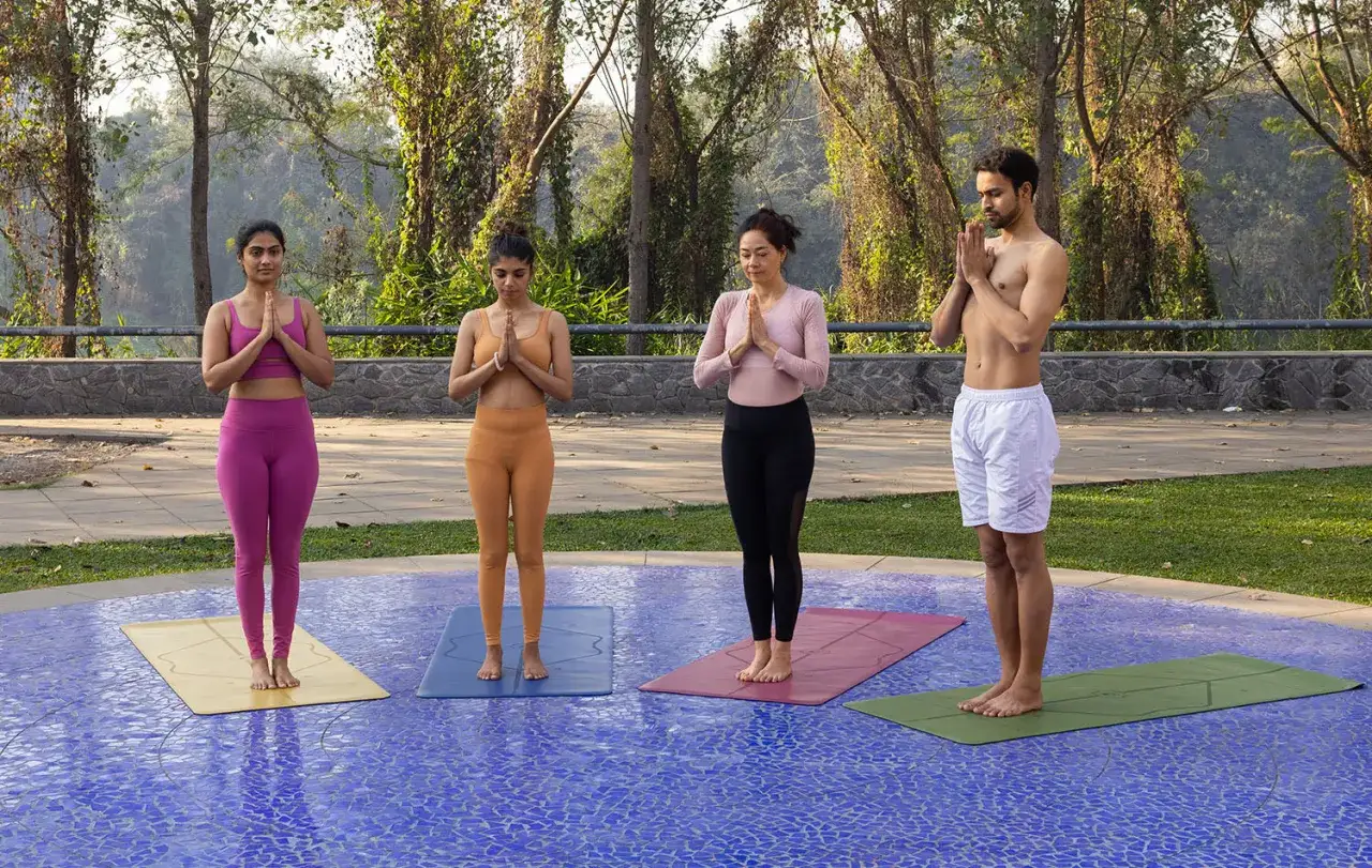 Vier Personen üben Hatha Yoga, 12 Grundstellungen, im Freien. Sie stehen auf Yogamatten mit gefalteten Händen vor der Brust.