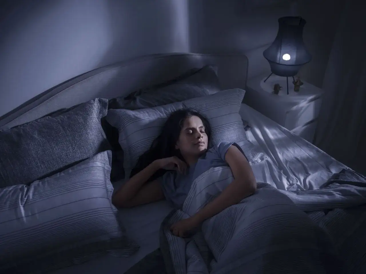 Eine Frau schläft tief und fest in einem gemütlichen Bett. Ihr Gesicht ist entspannt, und sie scheint einen leichten Schlaf zu genießen, während das sanfte Licht einer Nachttischlampe den Raum erhellt.