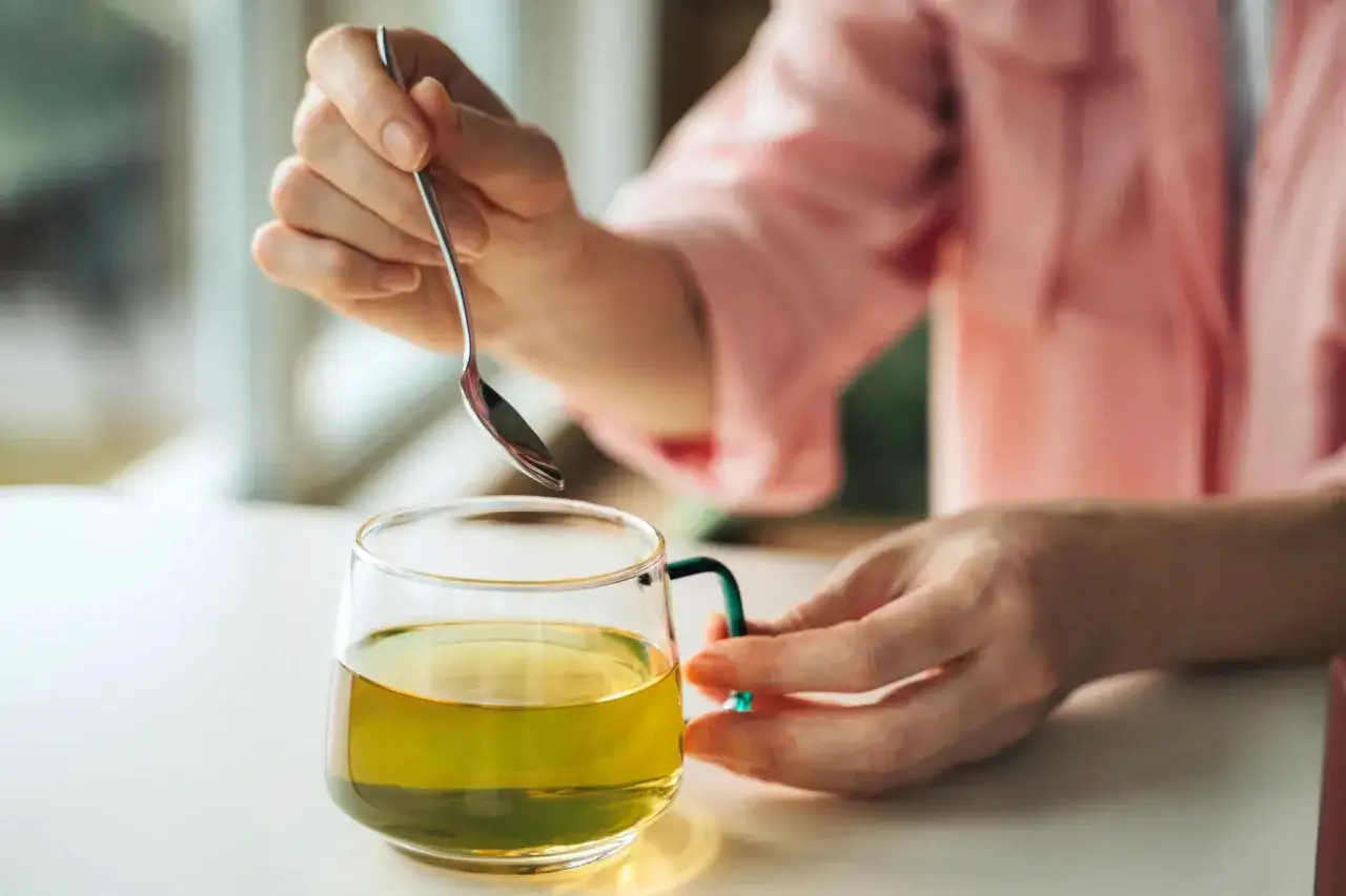 Eine Hand rührt mit einem Löffel in einer Tasse grünen Tees. Grüner Tee ist bekannt dafür, welcher Tee senkt Cholesterin und Blutdruck.