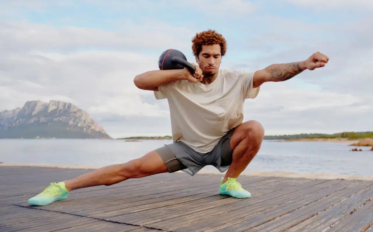 Mann trainiert mit Kettlebell am Meer, seitliche Ausfallschritte, ohne schuhe auf Holzplanken.